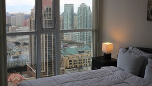 A hotel room with a large window offering a city view, a neatly made bed, and a bedside table with a lamp.