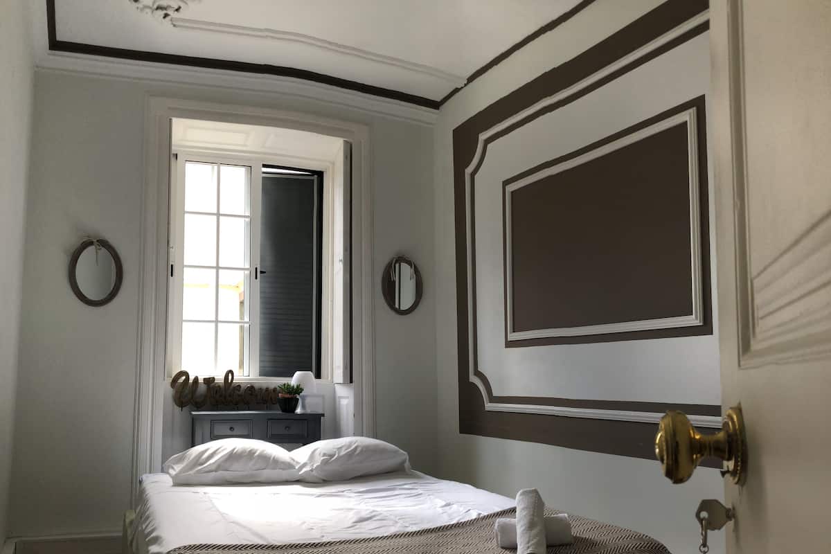 A bedroom with a bed, a window, a door, and a wall-mounted mirror.
