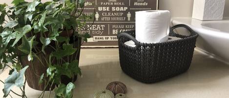 A bathroom counter with a sign listing bathroom rules, a potted plant, a basket with a towel, and two decorative shells.