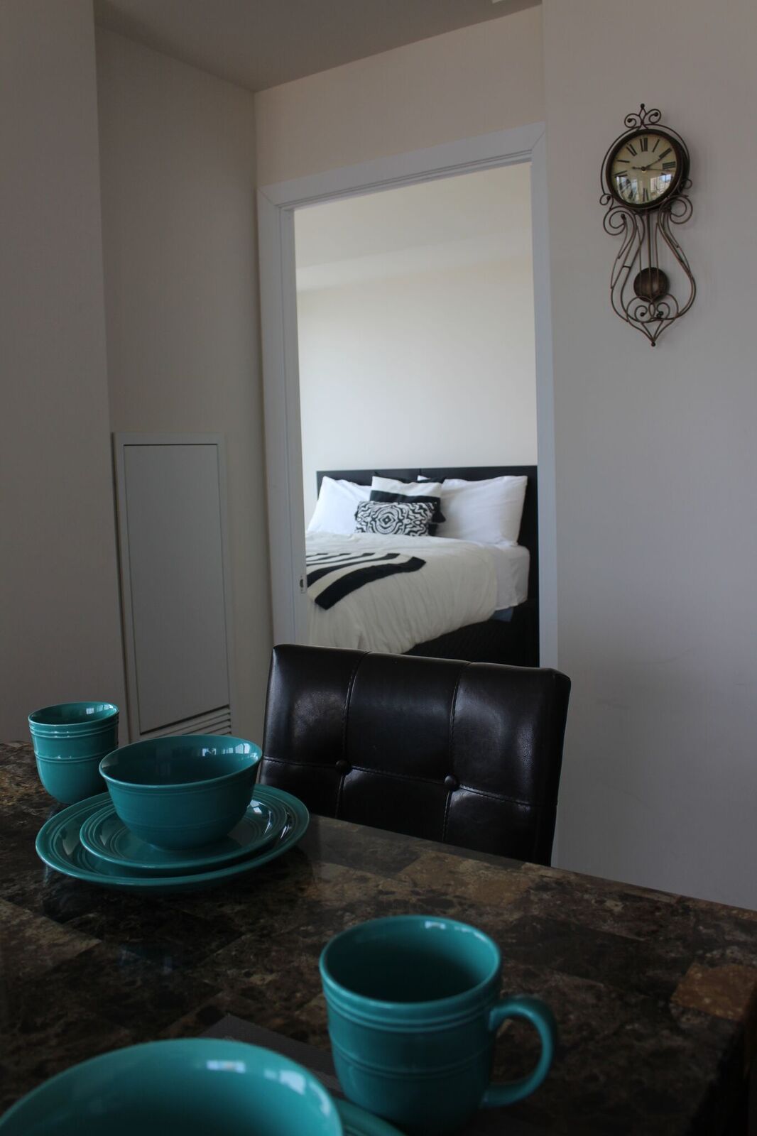 Une petite cuisine avec des bols de couleur turquoise et une tasse posée sur le comptoir, une chaise en cuir noir et, au fond, un lit agrémenté d’oreillers à motifs.