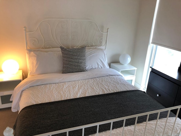 Un lit bien fait, avec une literie blanche et un plaid noir, une tête de lit en métal blanche, une table de chevet avec une lampe, et un petit objet rond blanc posé sur la table de chevet.