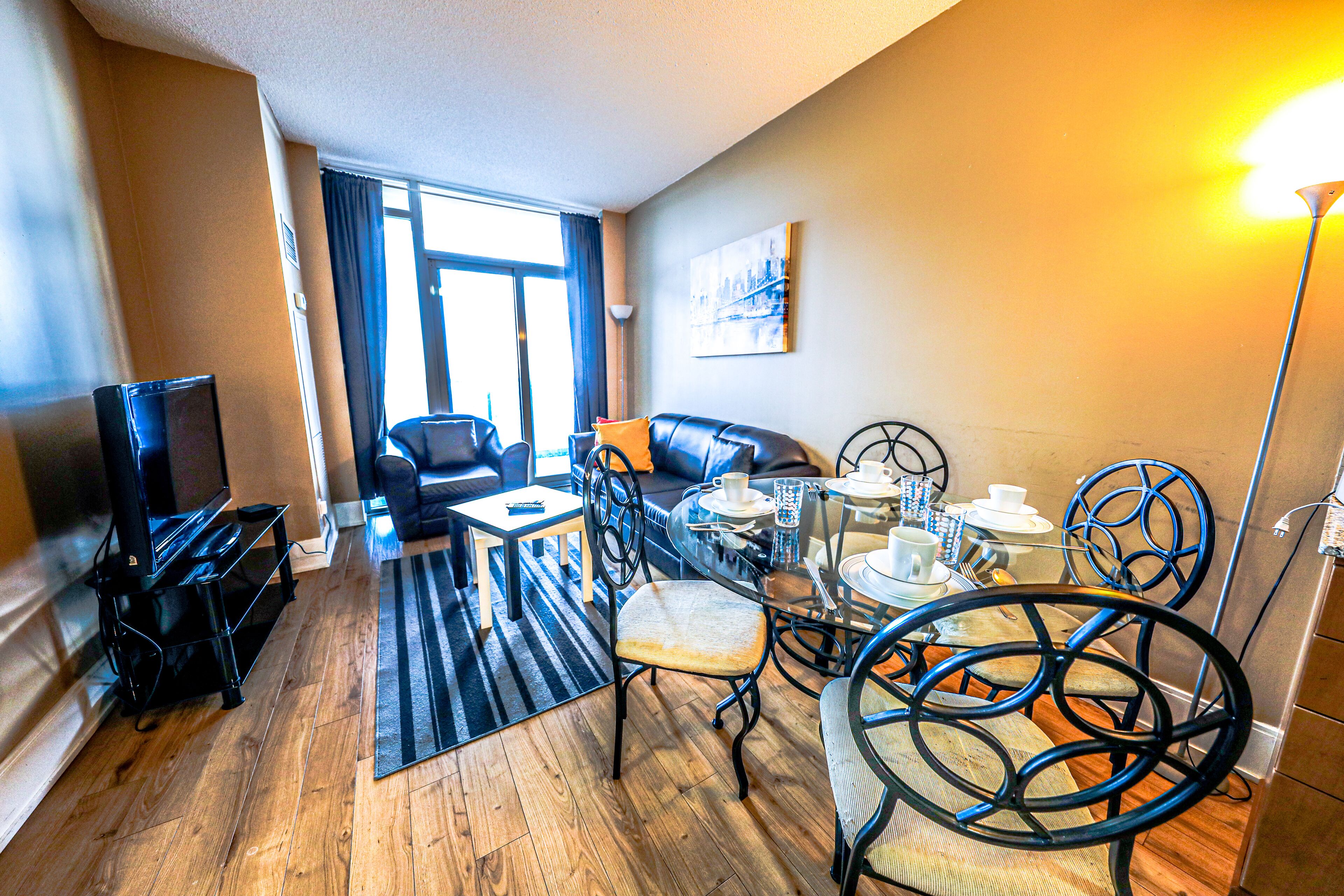 A hotel room with a wooden floor, a sofa, a dining table with chairs, a TV, and a painting on the wall.