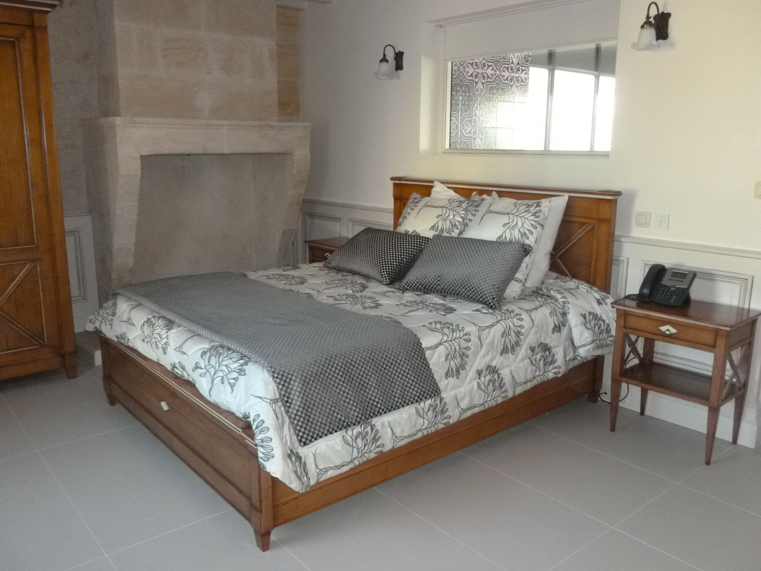 A bedroom with a wooden bed, a nightstand, a telephone, a mirror, and a stone fireplace.