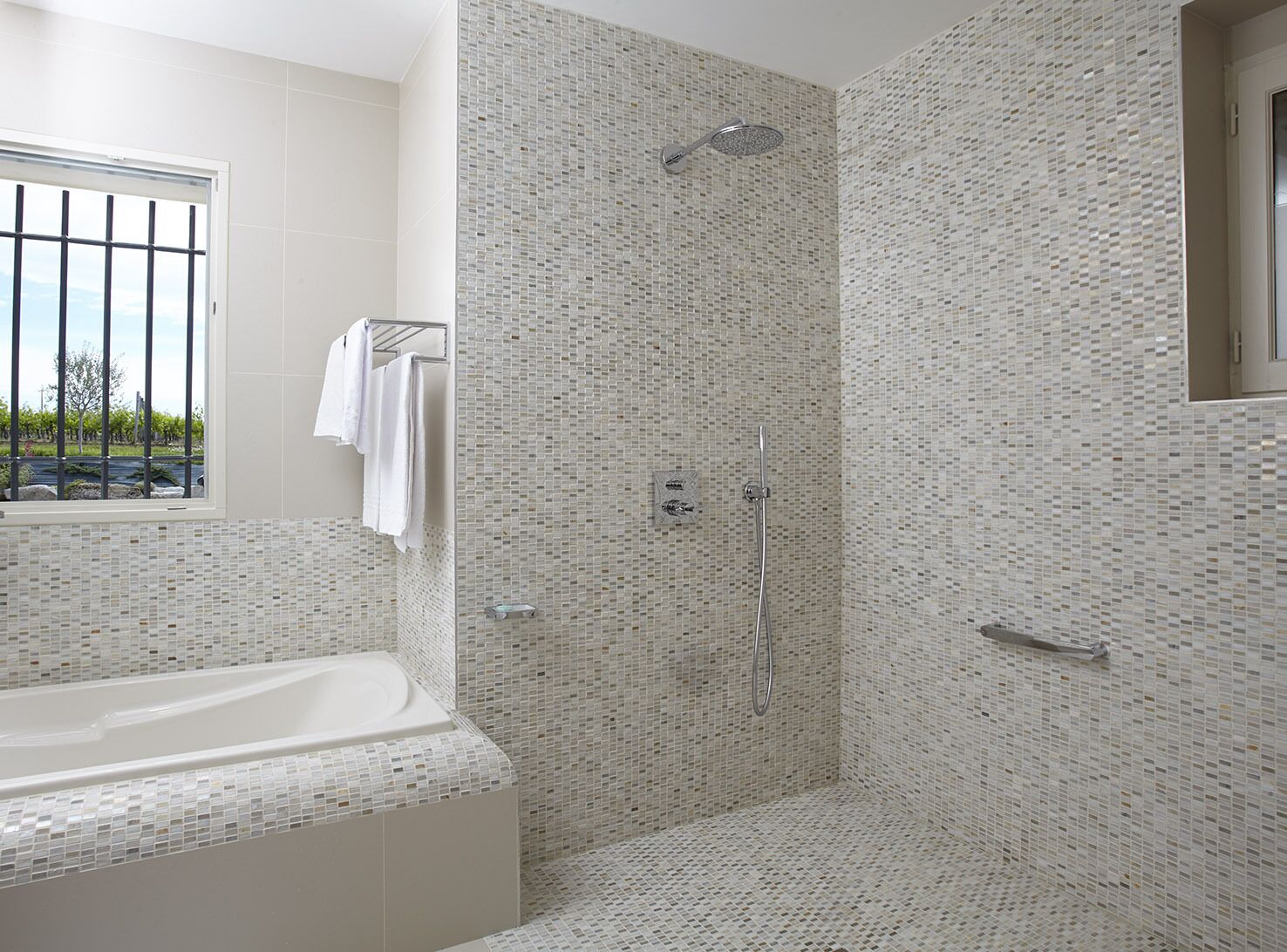 A modern bathroom with a mosaic tile shower wall, a freestanding bathtub, and a window with a view of greenery.