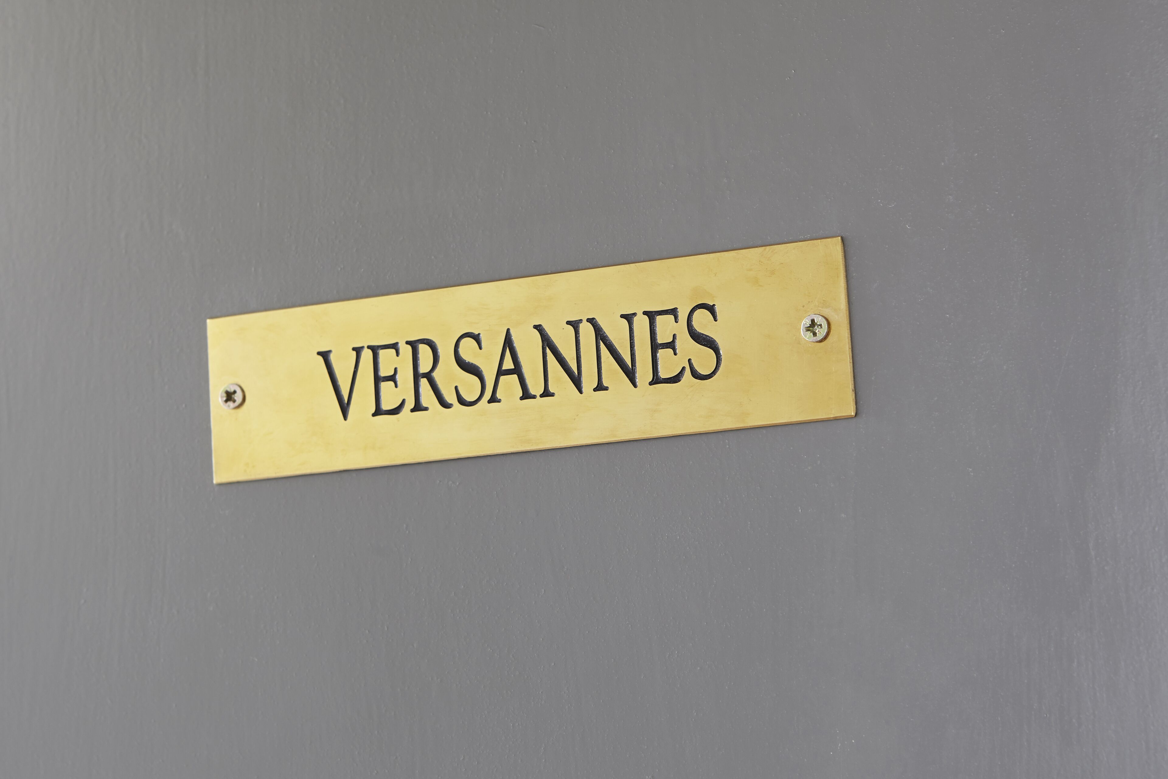 A brass nameplate with the inscription "VERSANNES" mounted on a grey wall.