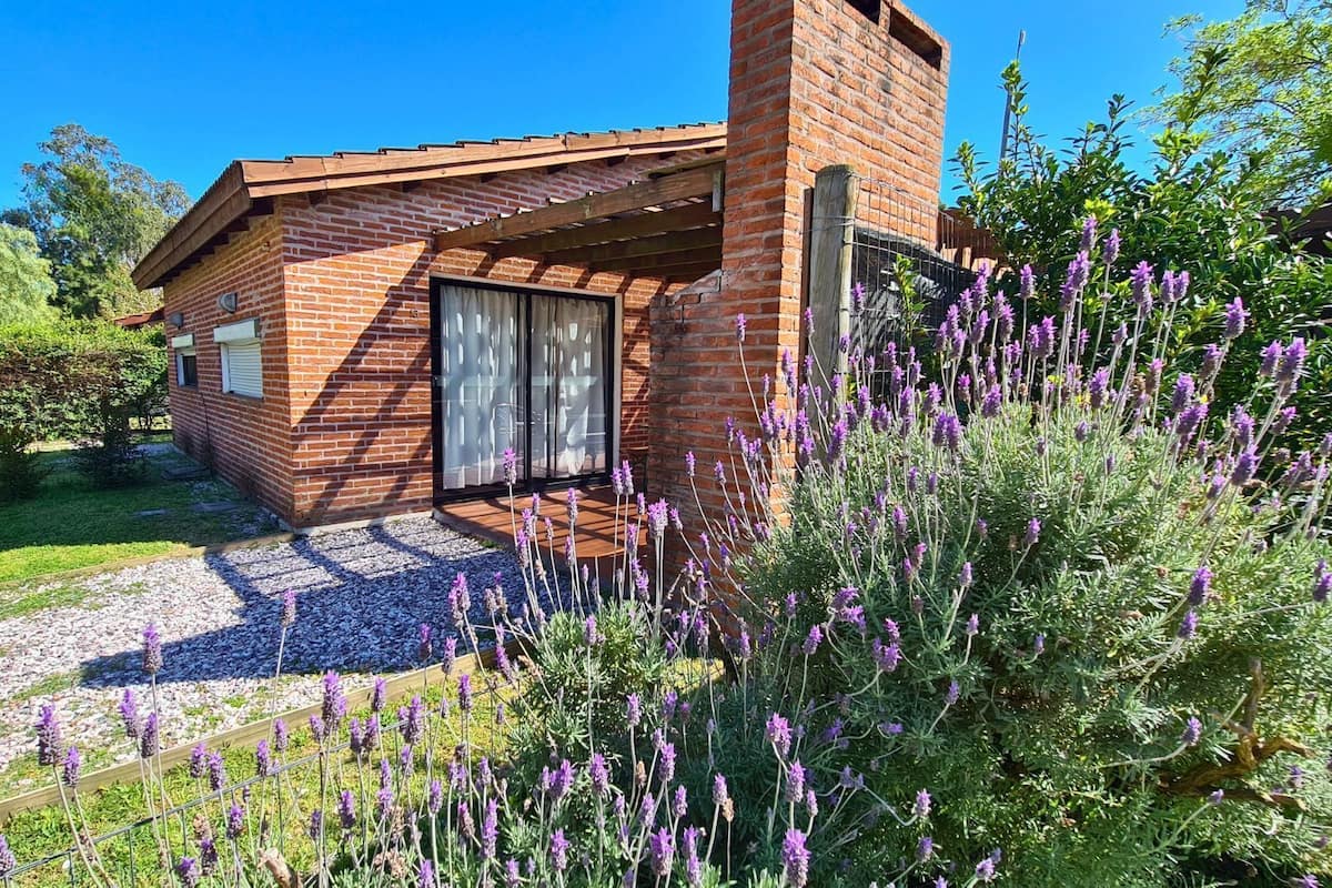 Una casa de ladrillo con terraza de madera y jardín lleno de flores moradas.