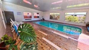 Une piscine intérieure dotée d’une terrasse en bois, de murs blancs et de fenêtres donnant sur un bâtiment en briques.