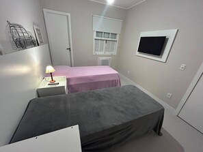 A small room with two beds, a desk with a lamp, a window with blinds, and a TV on the wall.