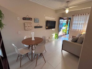 A modern living room with a round wooden table, white chairs, a beige sofa, and a flat-screen TV mounted on the wall. - Casa em Geribá com 2 quartos – acomoda 6 pessoas – Armação dos Búzios (Búzios)