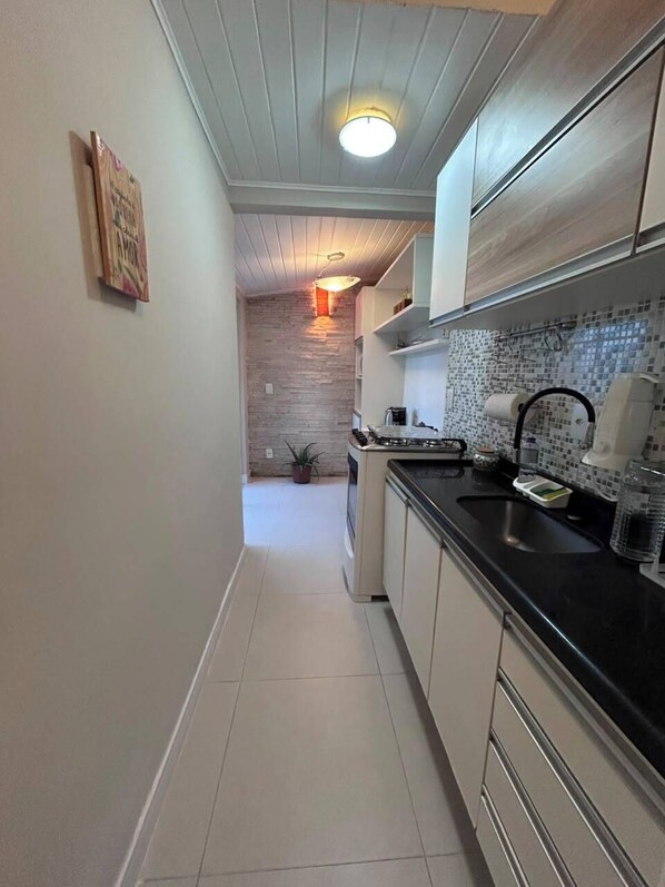 A narrow kitchen with white cabinets, a black countertop, and a tiled backsplash. - Casa em Geribá com 2 quartos – acomoda 6 pessoas – Armação dos Búzios (Búzios)