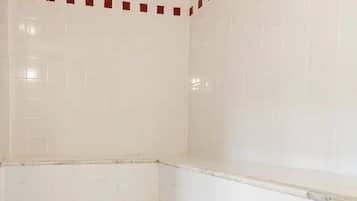 Casa de banho com paredes e chão em azulejo branco, zona de duche no canto e esgoto no chão.