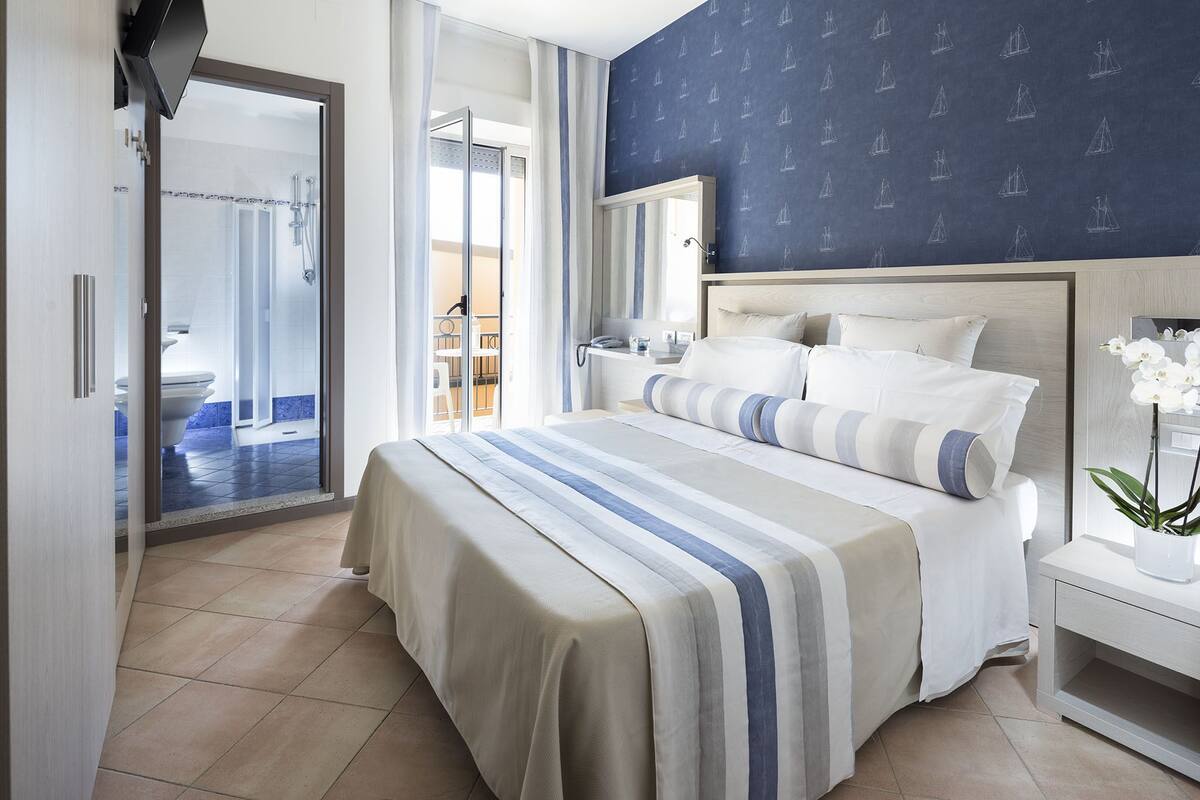 A hotel room with a large bed, a bathroom with a shower, and a view of the outside.