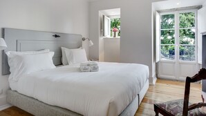 A neatly made bed with white linens and pillows, a bedside lamp, and a small window with a view of greenery.