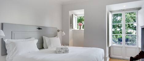 A neatly made bed with white linens and pillows, a bedside lamp, and a small window with a view of greenery.