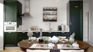 A modern kitchen with dark green cabinets, a white countertop, and a chandelier. There is a dining table set with plates, glasses, and a vase.