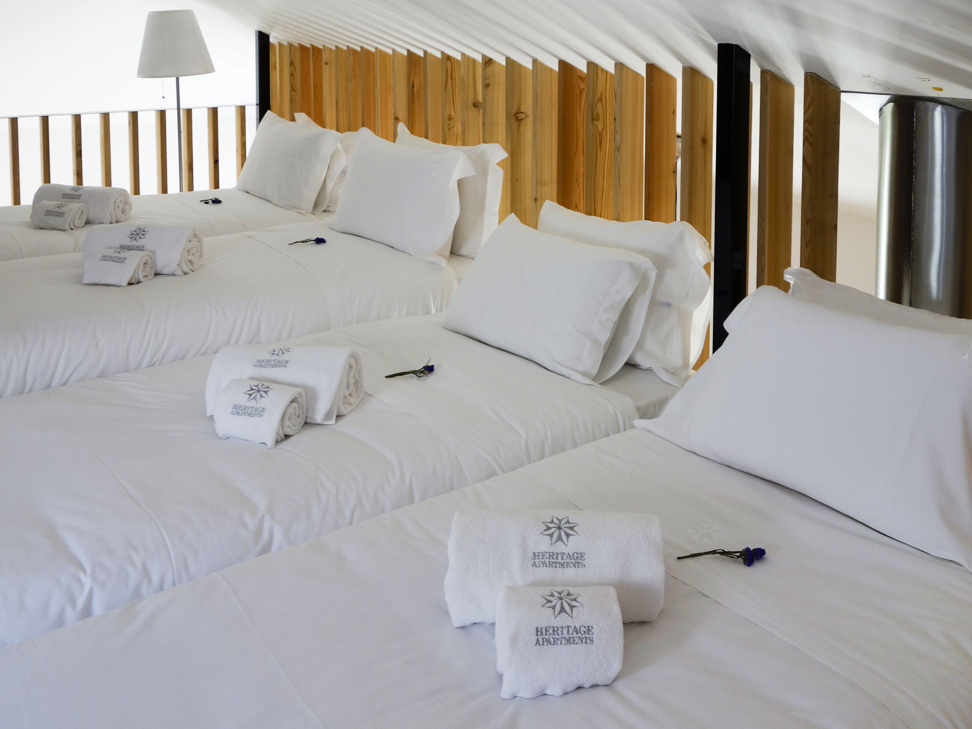 A hotel room with three beds, each with white linens and towels branded "Heritage Apartments."