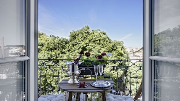 A balcony with a table set for two, a metal railing, and a view of a cityscape and greenery.