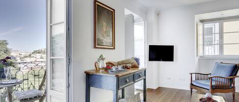 Une pièce spacieuse avec un balcon, une table en bois agrémentée d’objets décoratifs, un téléviseur fixé au mur et un fauteuil confortable.