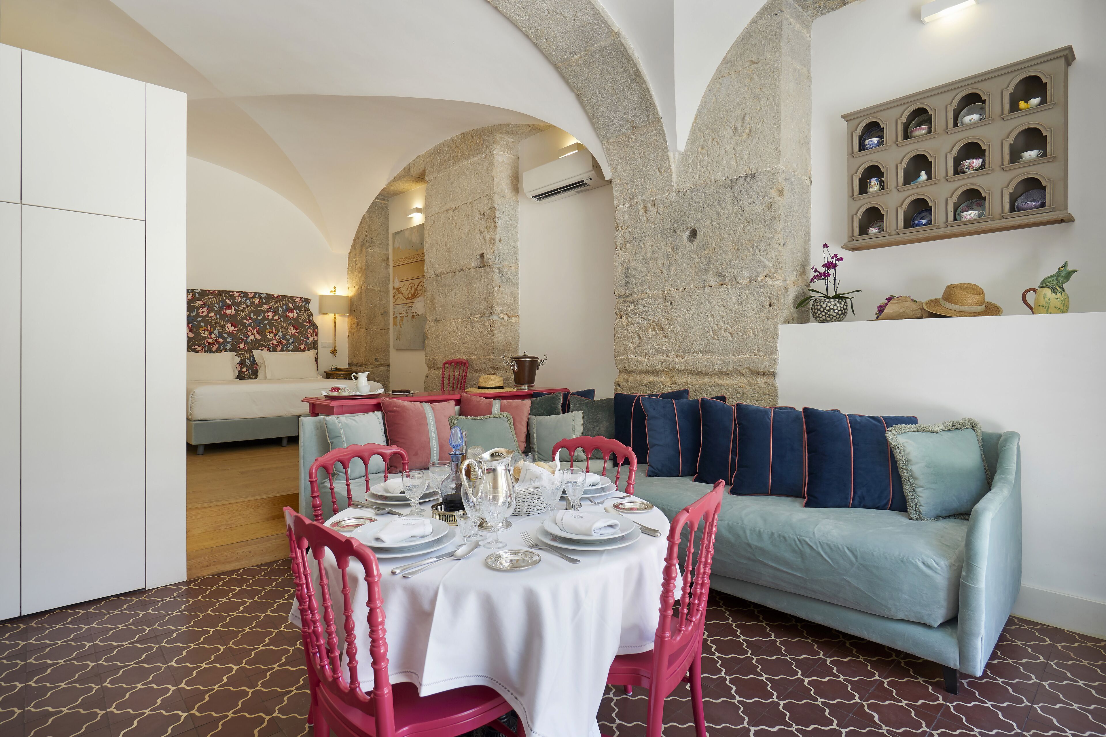 A dining area with a round table set for four, surrounded by pink chairs. A light blue sofa is against the wall, and a shelf with decorative items is above it.