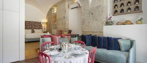 A dining area with a round table set for four, surrounded by pink chairs. A light blue sofa is against the wall, and a shelf with decorative items is above it.