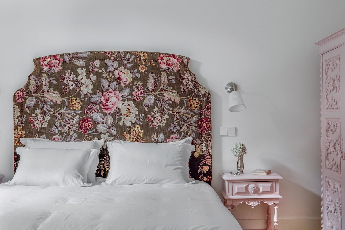 A neatly made bed with white linens, a floral headboard, a bedside table with a lamp, and a decorative cabinet.