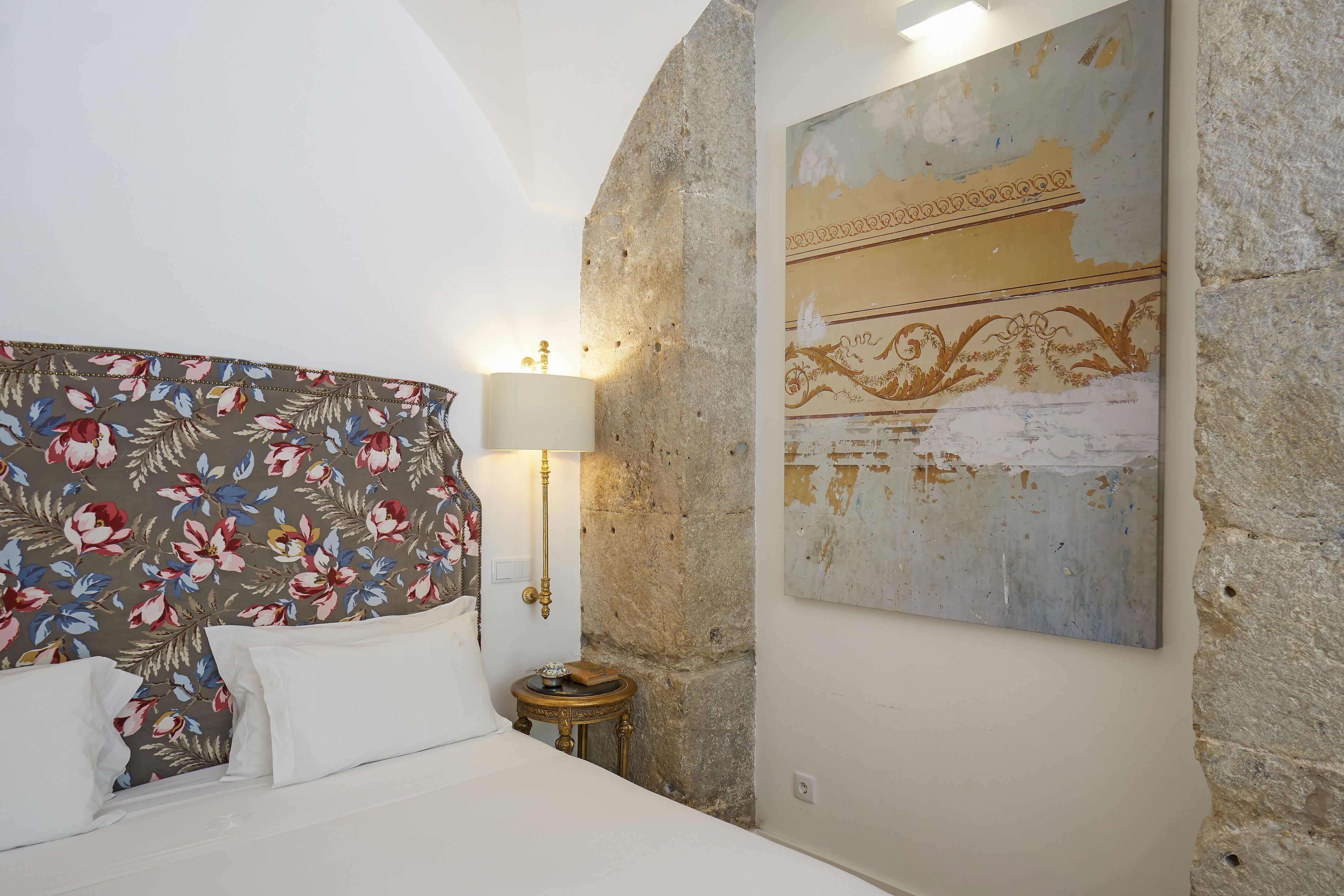 A bedroom with a floral headboard, a bedside table with a lamp, and a large decorative painting on the wall.