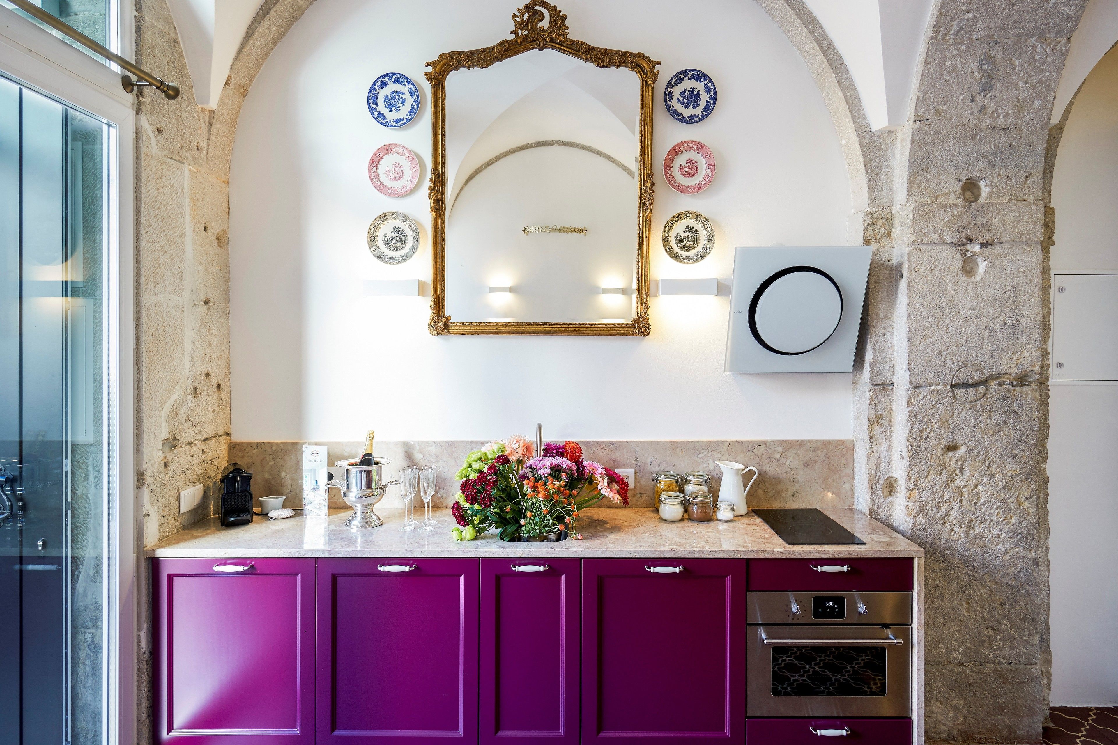 A kitchen with purple cabinets, a marble countertop, a decorative mirror, and a floral arrangement.