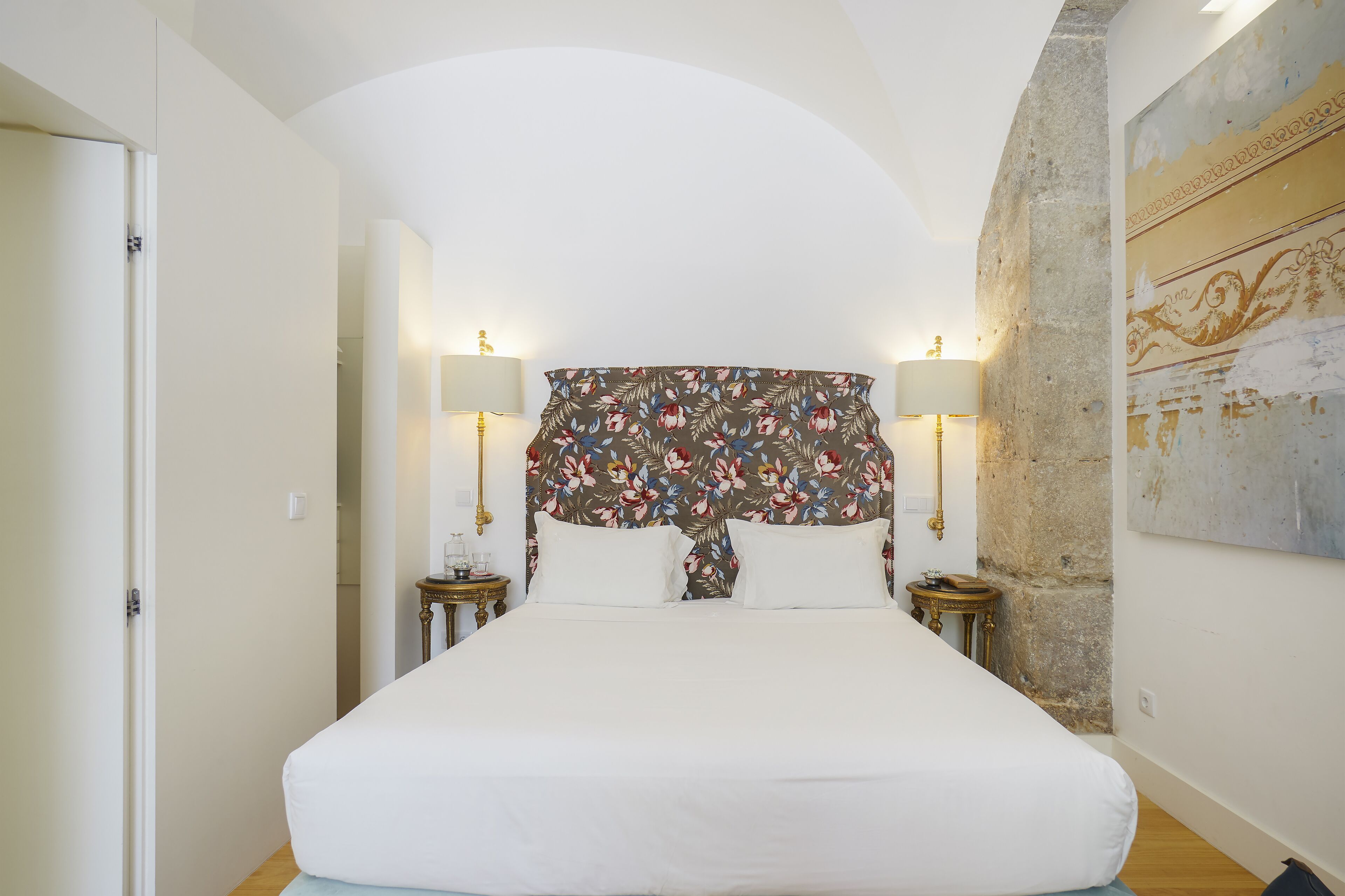 A bedroom with a large bed, two bedside tables with lamps, a floral headboard, and a stone wall.