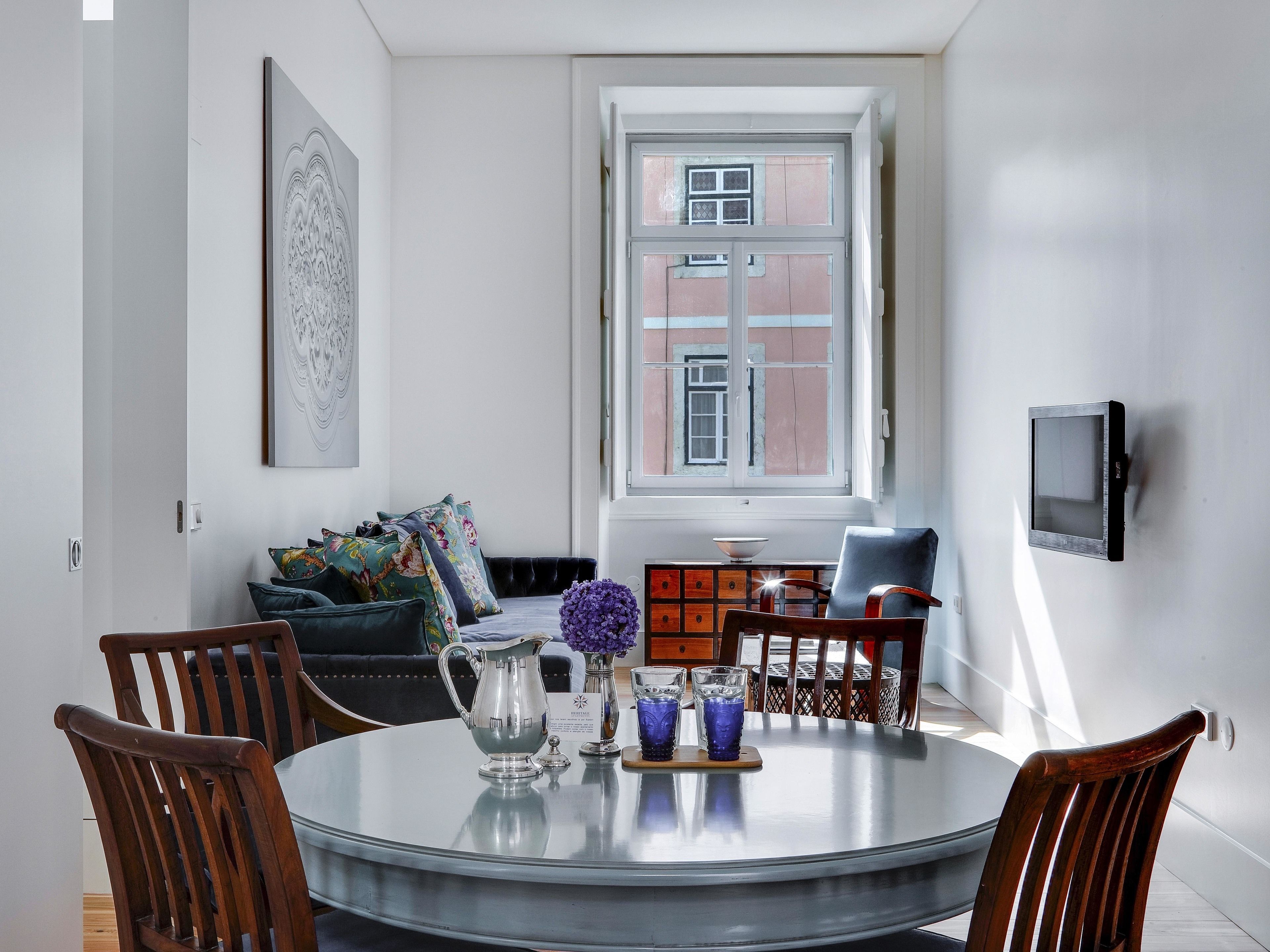 A dining area with a round glass table, wooden chairs, a silver pitcher, and blue glassware. A sofa with colorful cushions is against the wall, and a window with a view of buildings is visible.