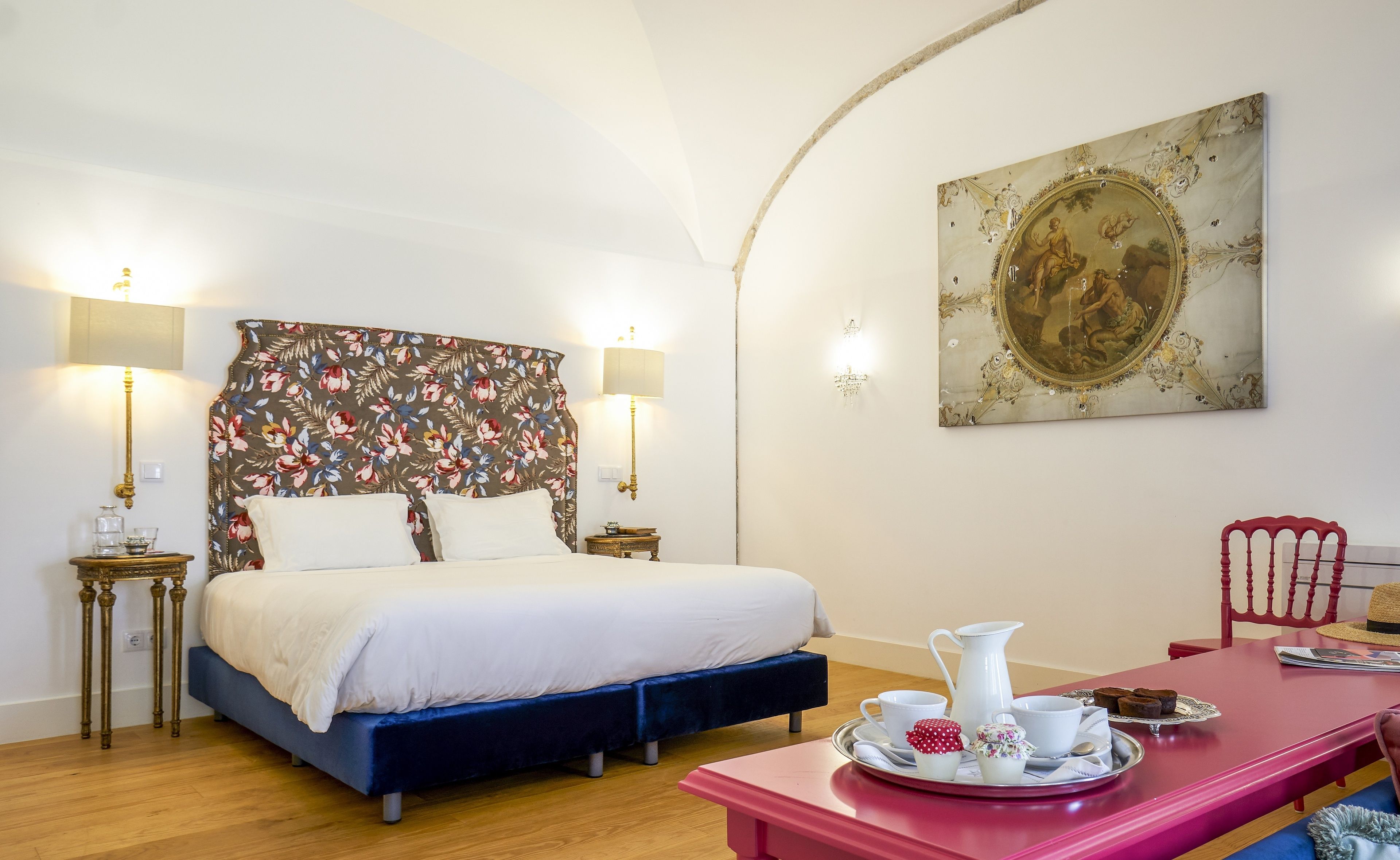 A bedroom with a bed, bedside lamps, a decorative headboard, a dining table with a tray of items, and a wall painting.