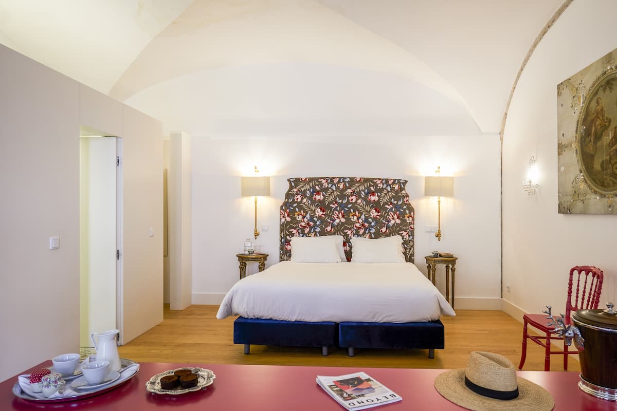 A bedroom with a bed, bedside tables, a red chair, a wooden side table, and a tray with cups and a plate.
