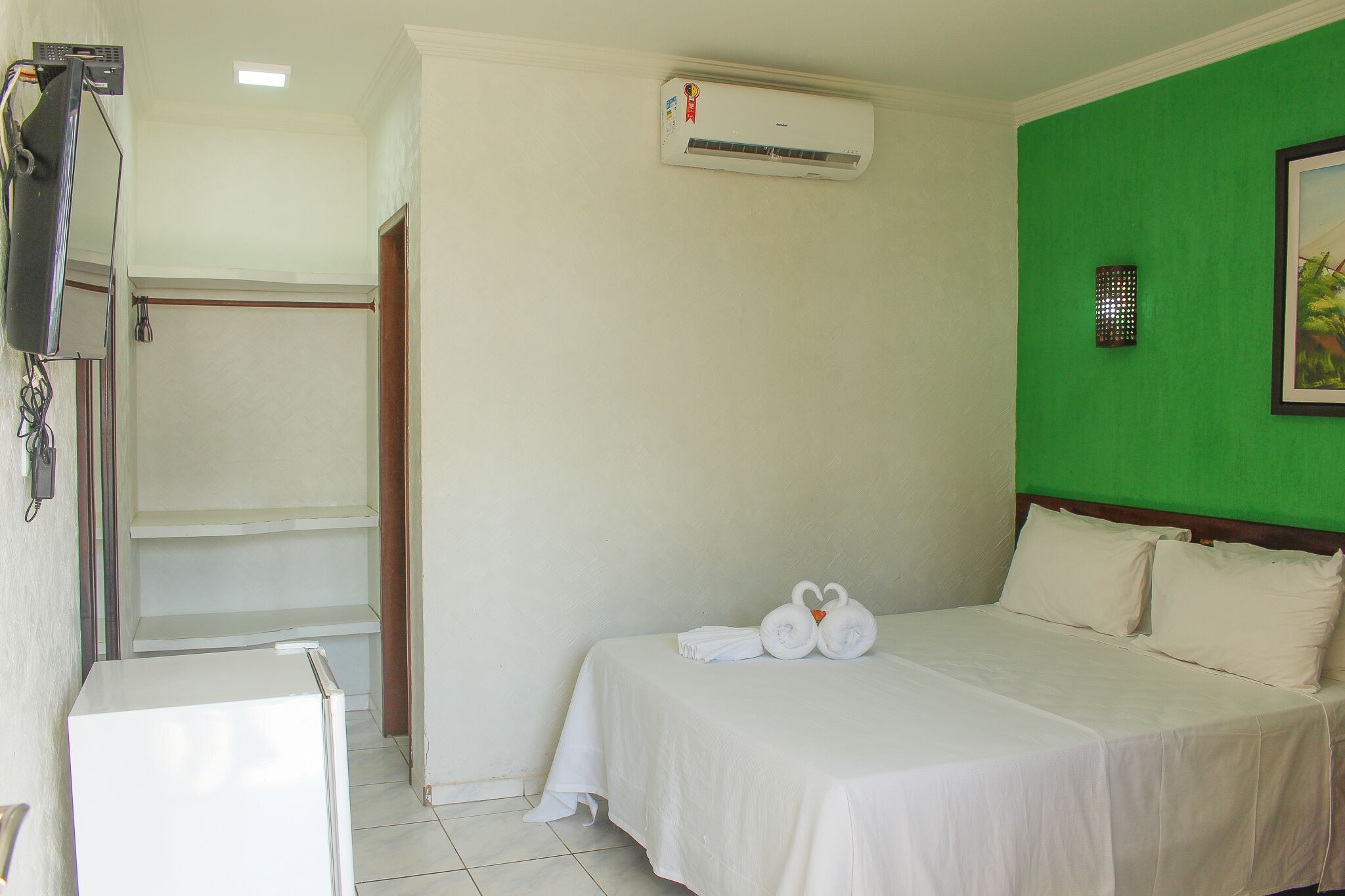 Habitación de hotel con cama, refrigerador blanco pequeño, aire acondicionado de pared y una pared de acento verde.