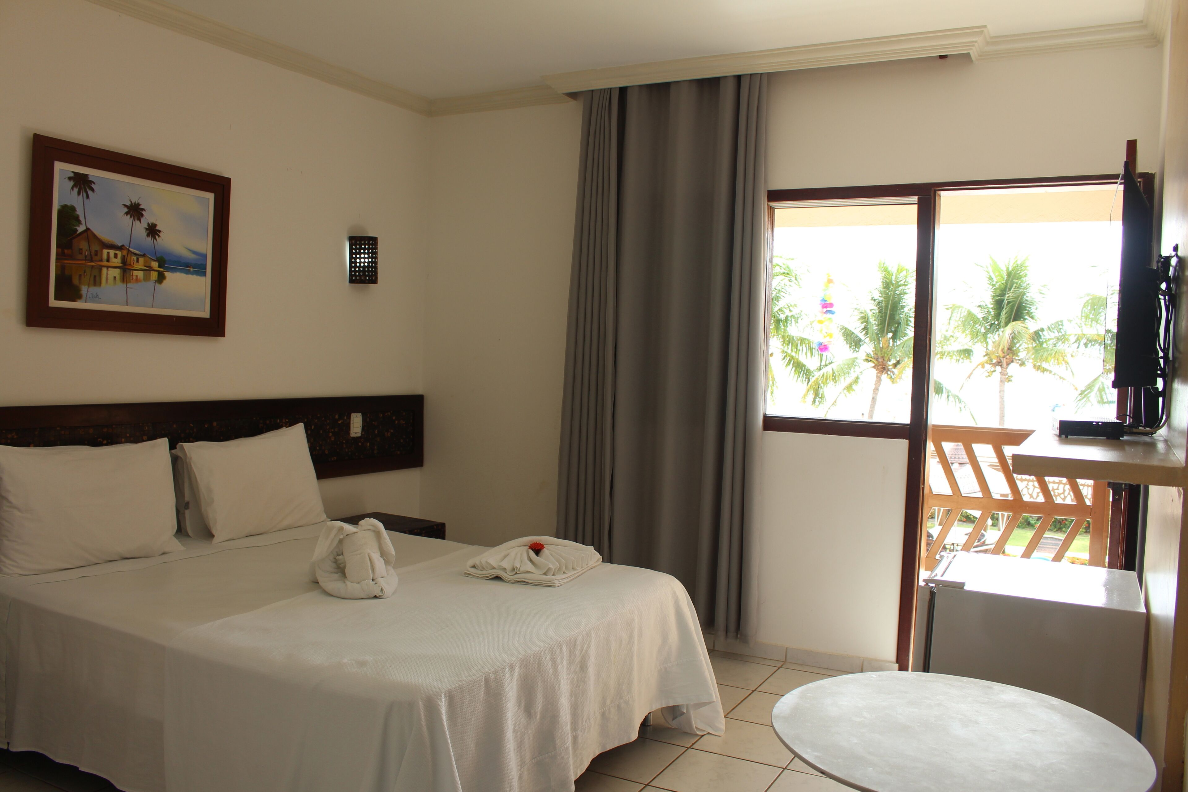 A hotel room with a bed, a small table, a TV, a painting, and a window with a view of palm trees.