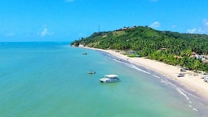 Une plage côtière aux eaux turquoise et limpides, un bateau près du rivage, et une colline couverte de végétation et parsemée de bâtiments.