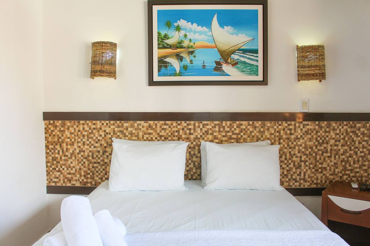 A hotel room with a bed, two pillows, a headboard with a pixelated pattern, and a framed painting of a tropical scene above the bed.