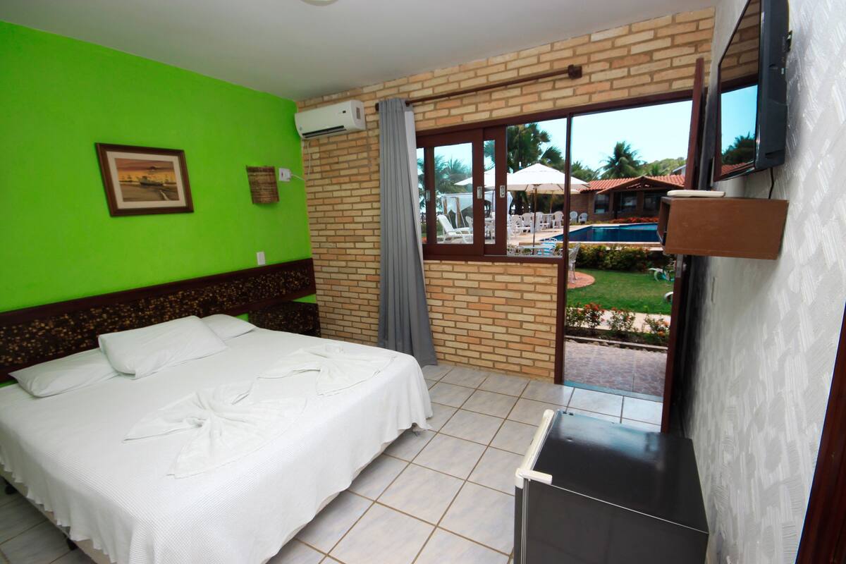 A hotel room with a bed, a TV, a window with a view of a pool and greenery, and a brick wall.