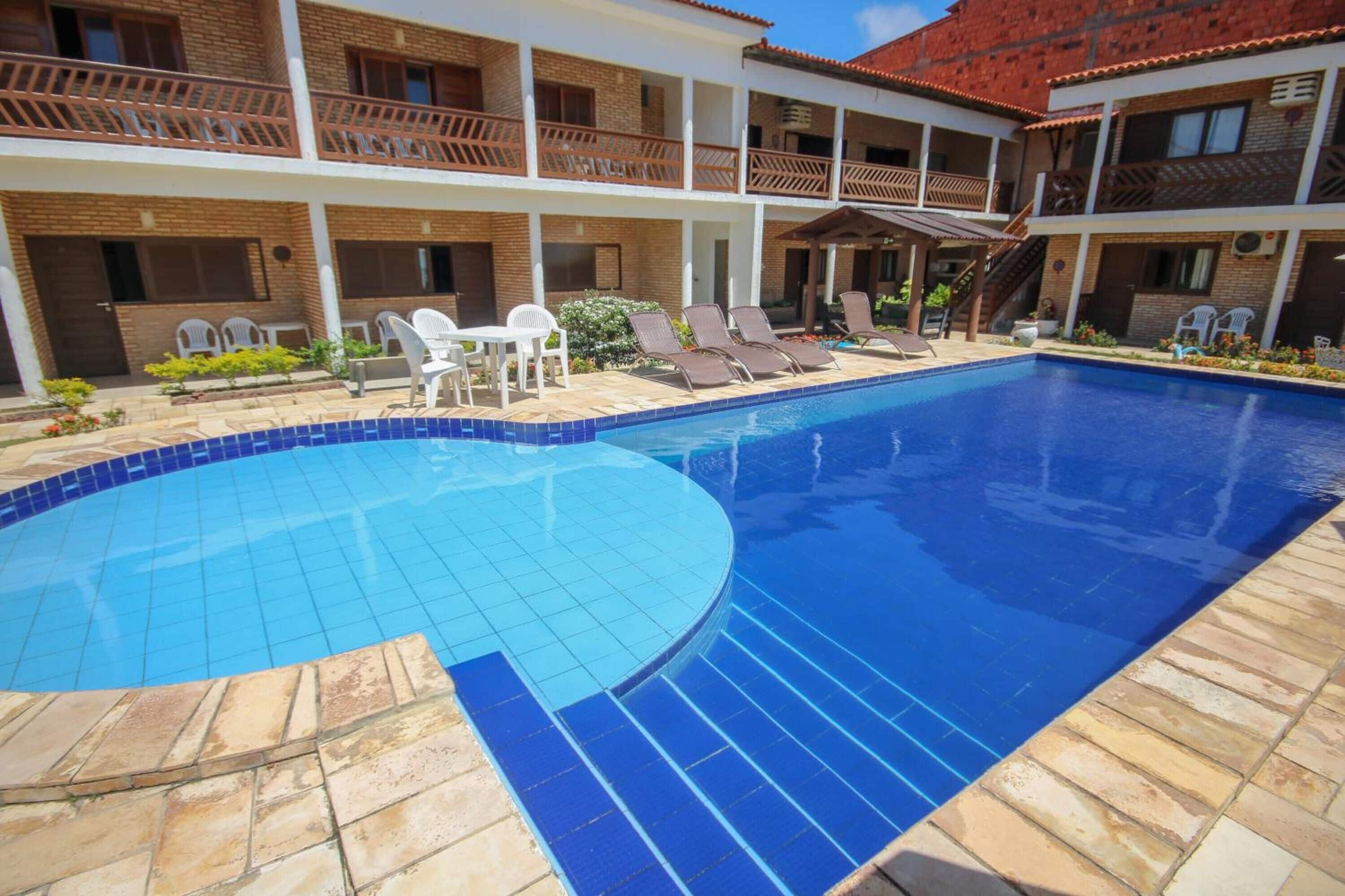 A swimming pool with tiled edges, surrounded by lounge chairs and a two-story building with balconies.