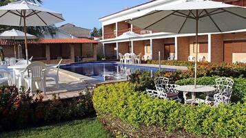 A pool area with white chairs and umbrellas, surrounded by well-maintained hedges and a brick building with balconies.
