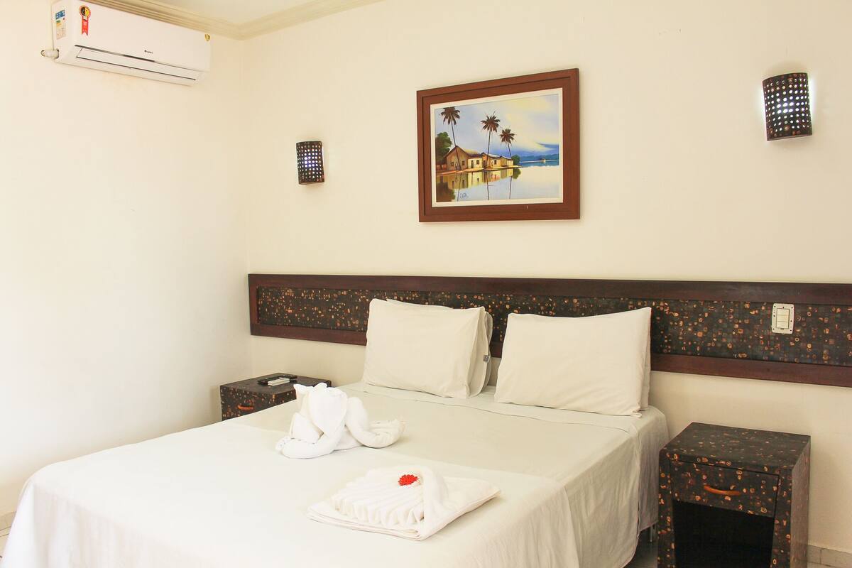 A hotel room with a bed, a painting, an air conditioner, and a nightstand.