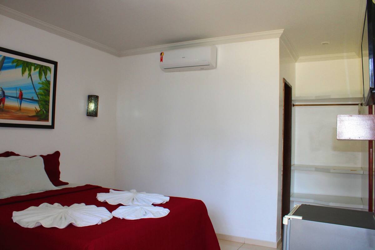 A hotel room with a bed, a painting, an air conditioner, and a minibar.