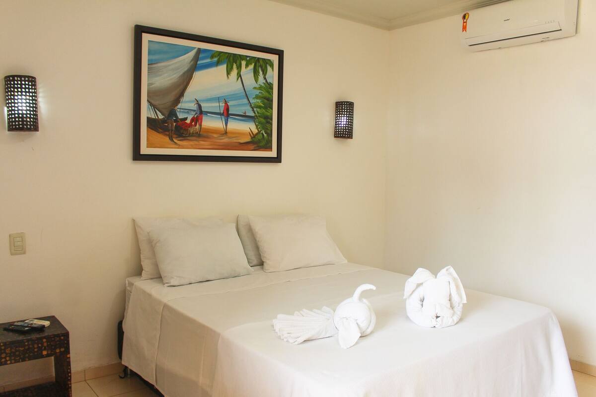 A neatly made bed with white linens and two towels shaped like animals, a painting of a beach scene above the bed, and two wall-mounted light fixtures.