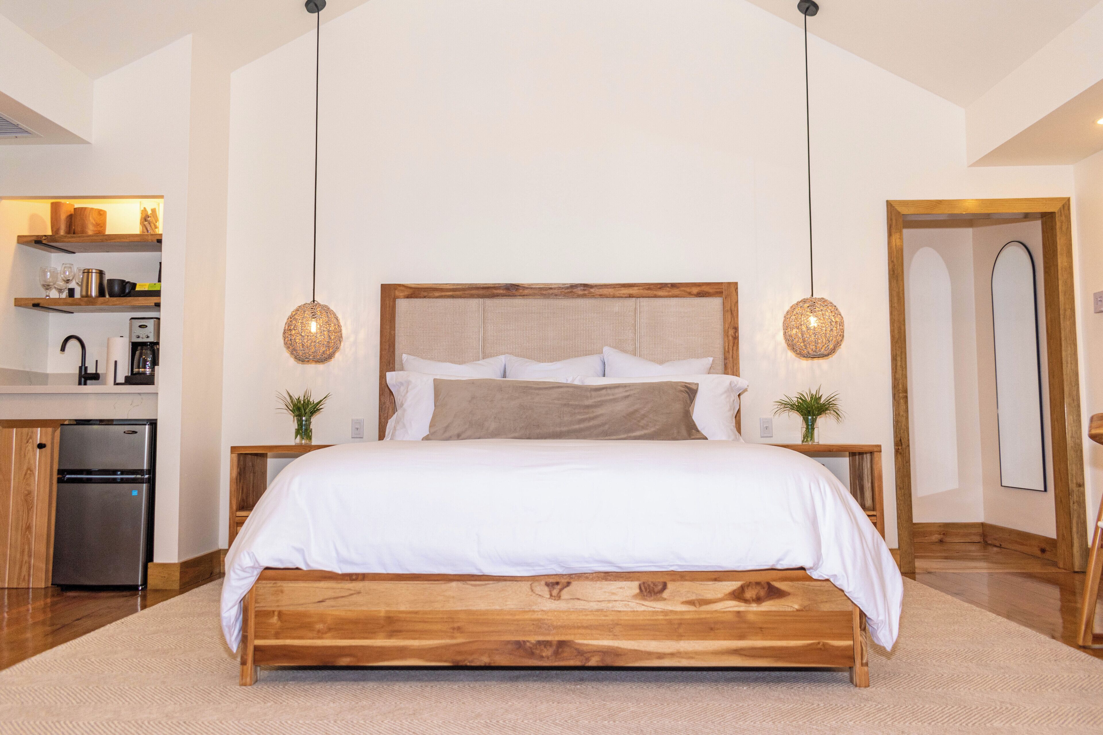 Un dormitorio con cama de madera, dos mesitas de noche, una mesita auxiliar, una lámpara colgante y una puerta de madera.