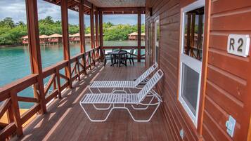 Terraza de madera con sillones y una mesa, con vista a un cuerpo de agua, edificios y follaje.