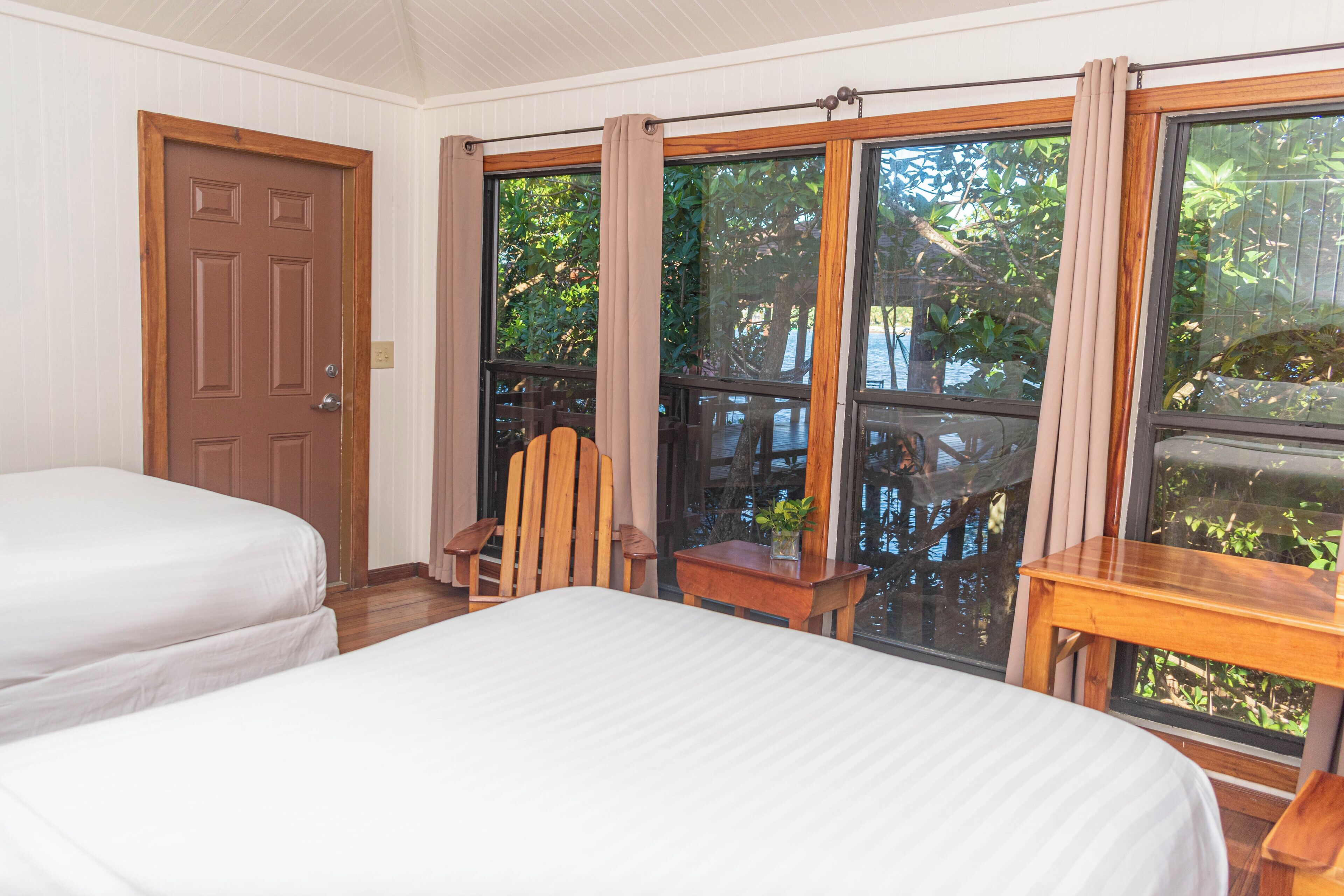 Una habitación con dos camas, una silla de madera, una mesita pequeña y ventanas grandes con cortinas.