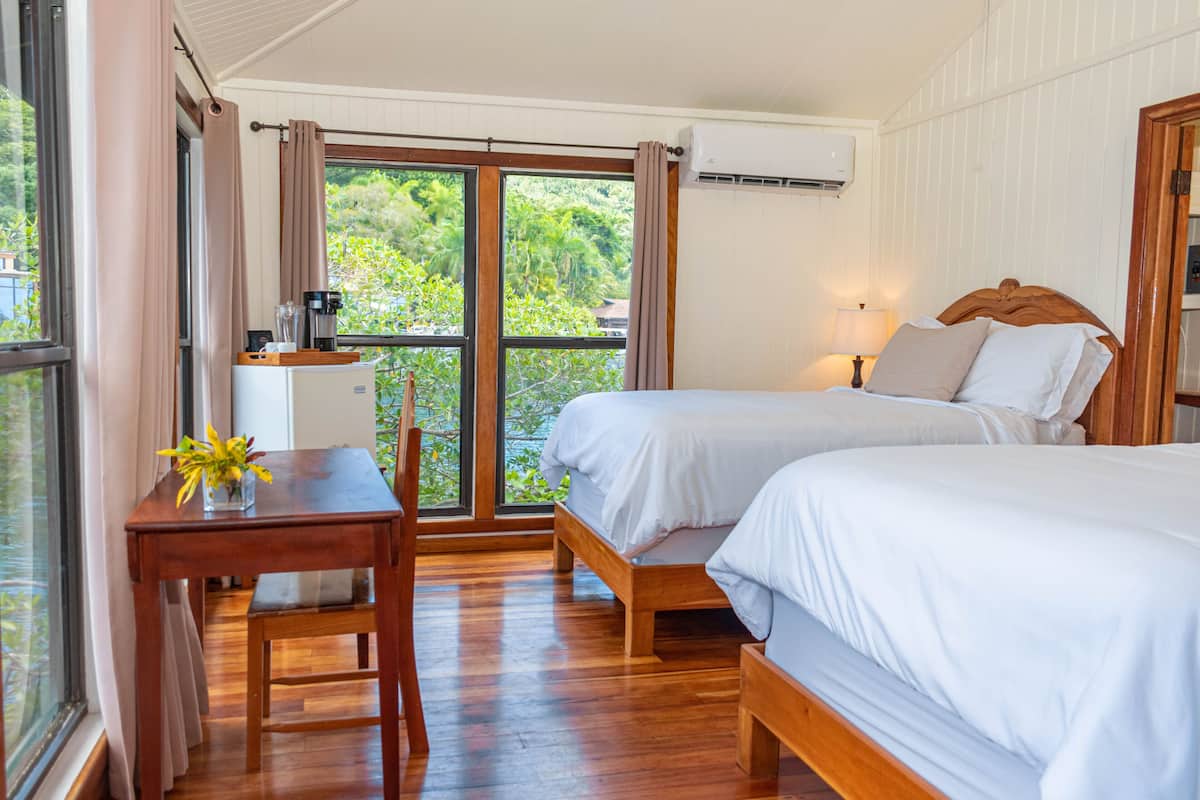 A bedroom with two beds, a wooden desk, a coffee maker, and a view of greenery through a large window.