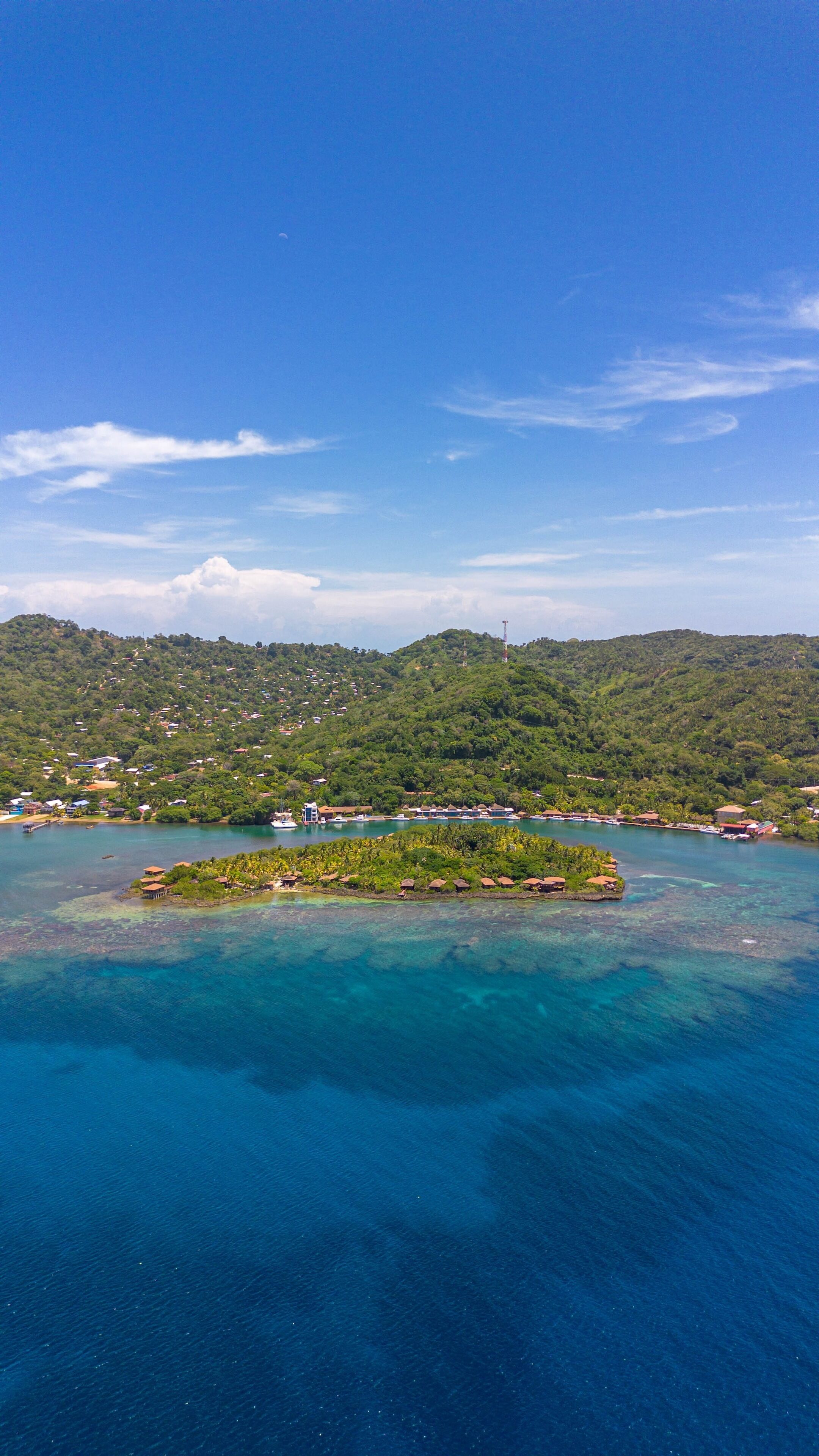 An aerial view of a coastal area with a small island featuring a cluster of buildings, surrounded by clear blue waters and a backdrop of green hills.