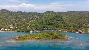 An aerial view of a coastal area with a small island featuring a cluster of buildings, surrounded by clear blue waters and a backdrop of green hills.