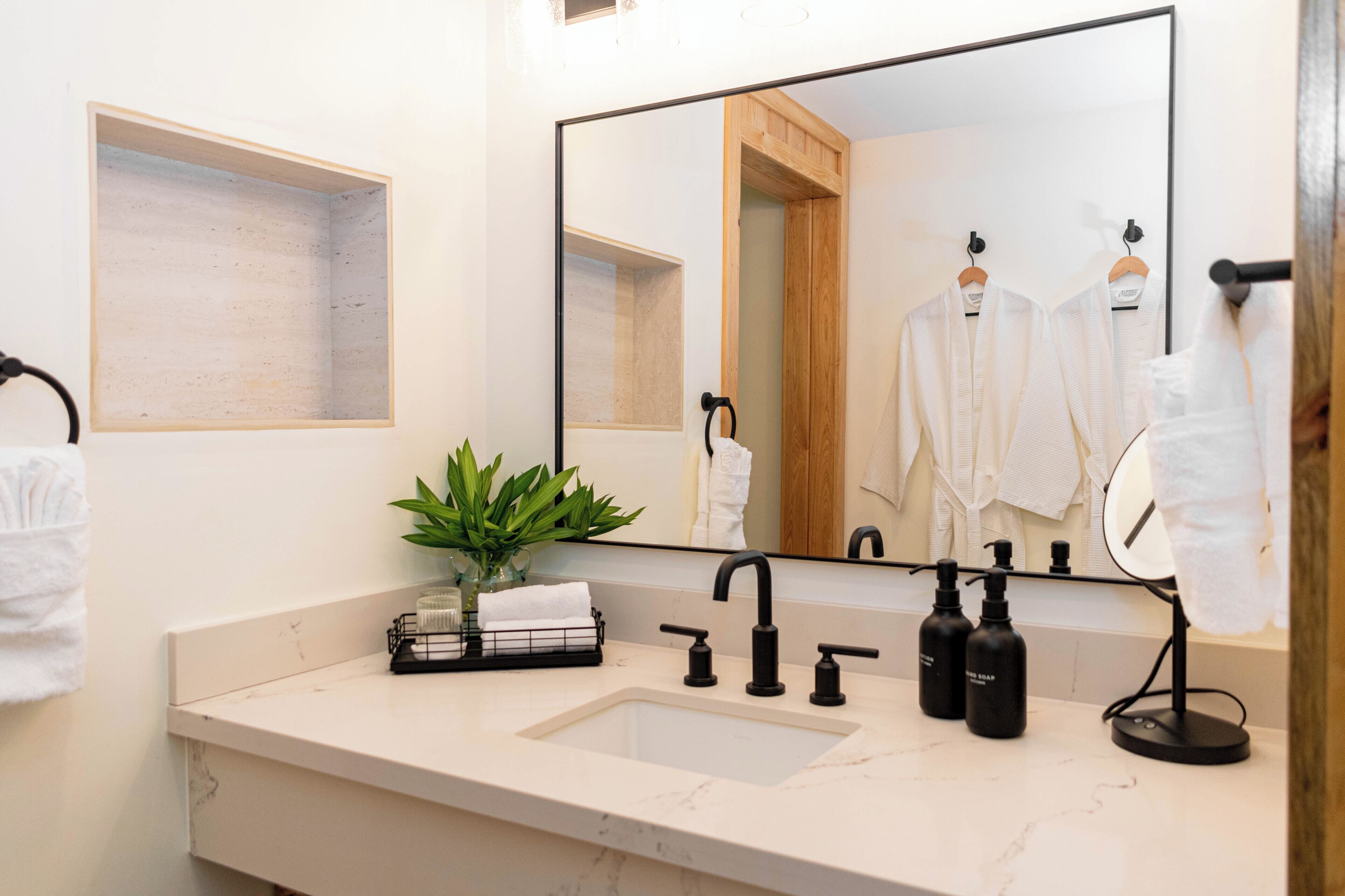 Un baño moderno con un espejo grande, un lavamanos con grifos negros, una planta pequeña y un perchero con toallas blancas.