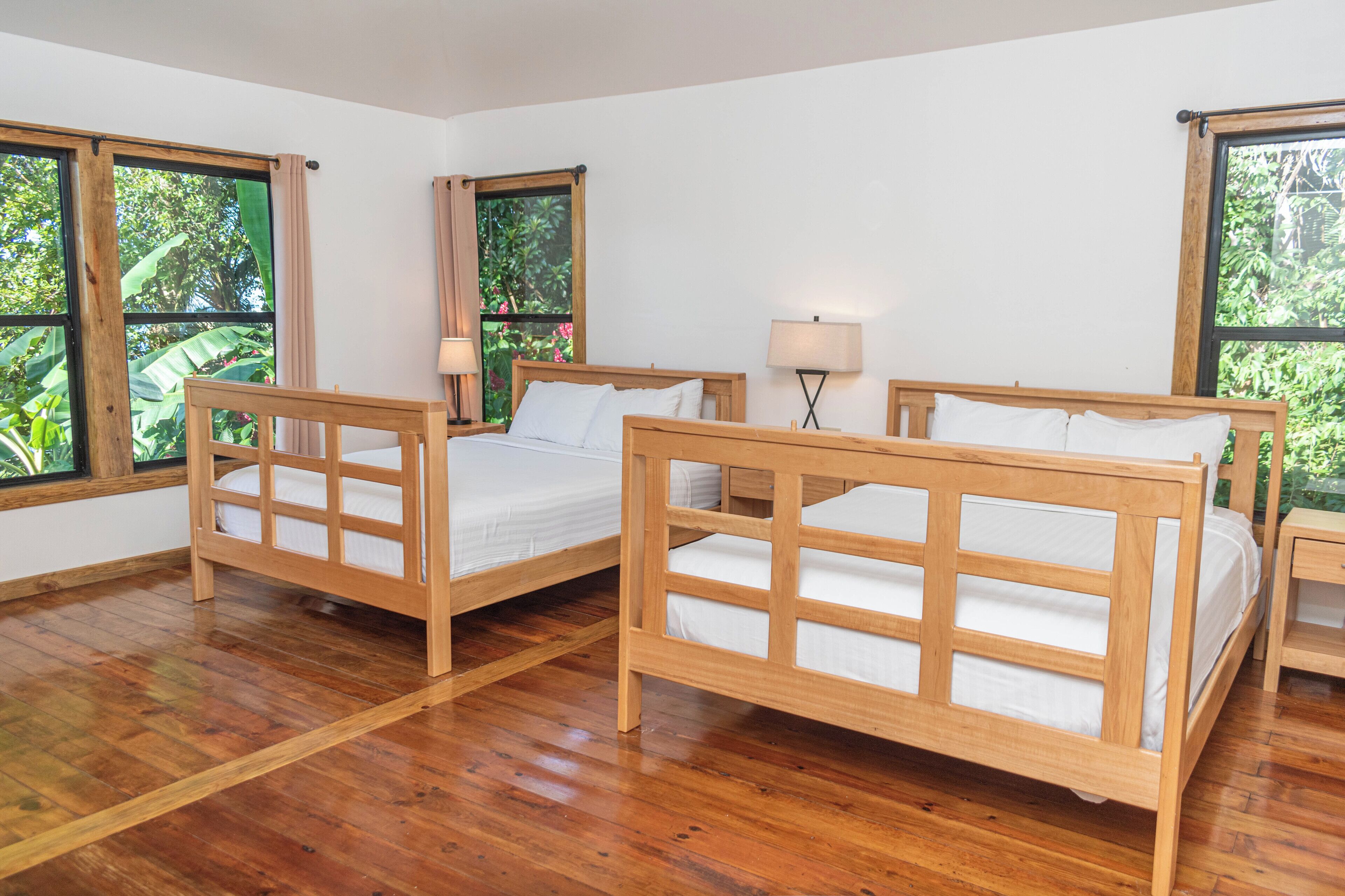 Une chambre avec deux lits en bois, une table de chevet avec une lampe et de grandes fenêtres donnant sur un paysage verdoyant.