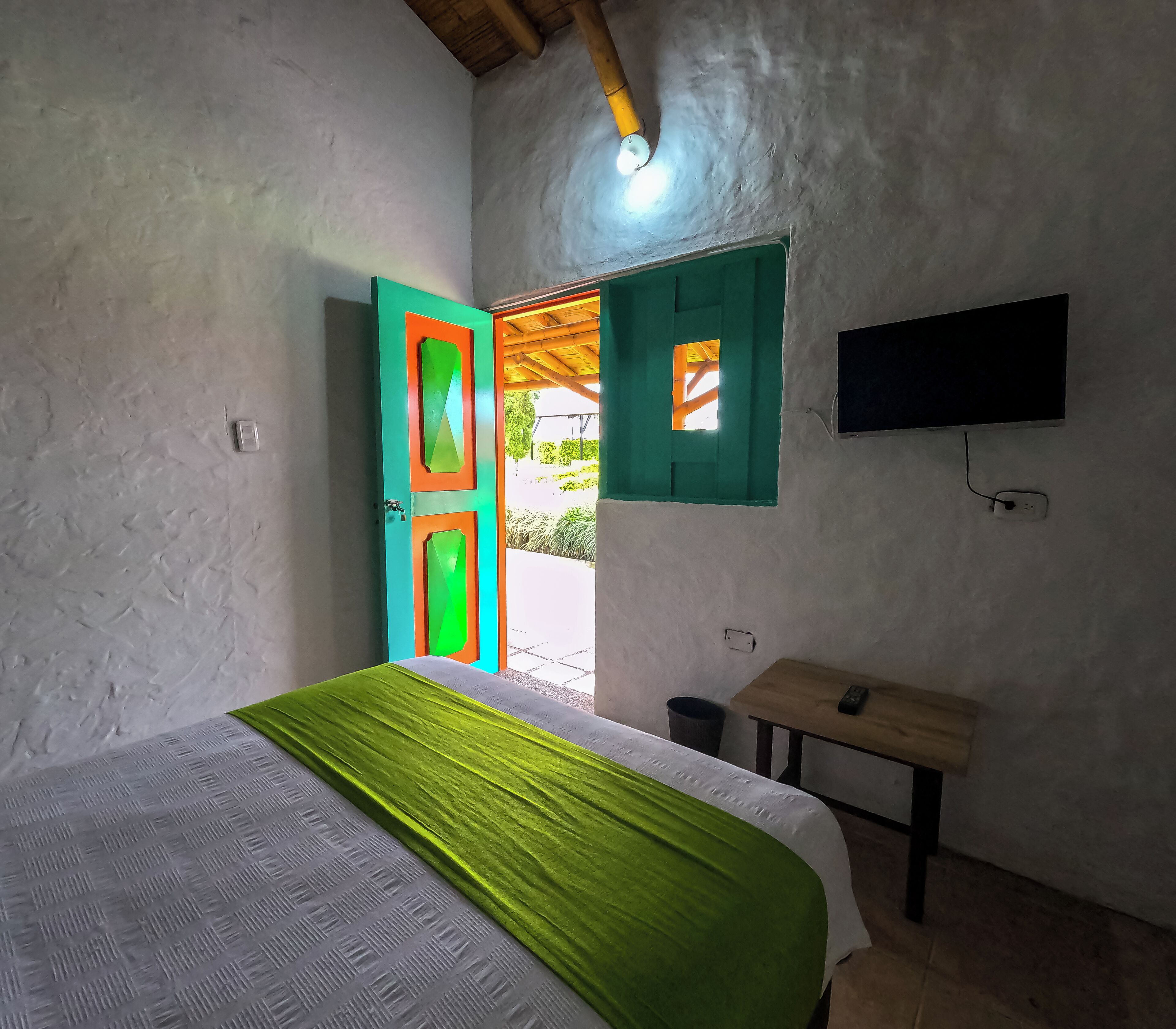 Une chambre avec un lit, une porte verte, une petite table, un téléviseur et une fenêtre donnant sur un paysage verdoyant.